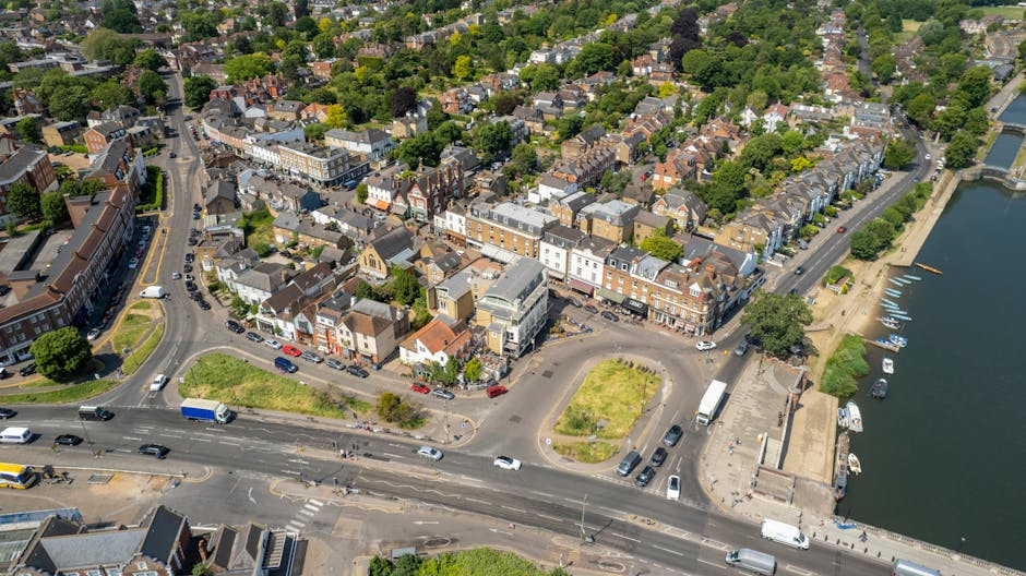 Aerial view of a town centre in Kingston upon Thames, showing a variety of buildings including shops, offices, and residential properties with a river on the right side of the image. The scene captures a busy street with cars, vans, and trucks parked along the side and moving through the intersection, highlighting typical urban traffic flow. In the foreground, a section of road is visible with a loading area adjacent to a parking lot containing several parked vehicles, some of which are being loaded or unloaded. The street features sidewalks, small green patches, and trees lining the sides, contributing to the overall landscape of the town. The riverbank on the right includes a promenade with boats docked along the quay, and a small pier with boats moored to it. This context of urban infrastructure supports the process of home relocation and furniture transport, as carried out by companies like Removals Kingston upon Thames during packing, loading, and transportation of belongings in Kingston upon Thames.