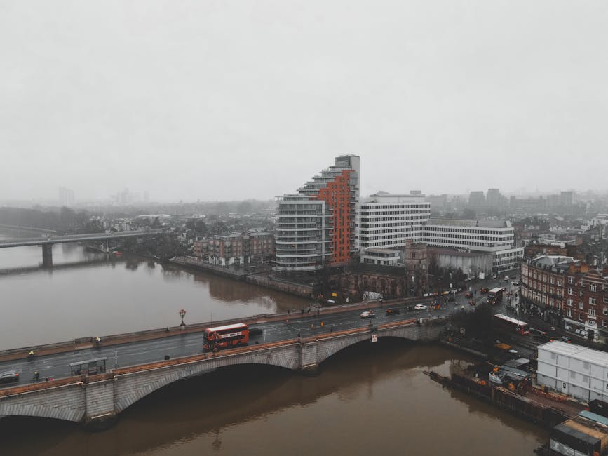 Aerial view of a multi-storey modern residential building with curved balconies, situated next to a river in Kingston upon Thames. The riverbanks are lined with brick and stone buildings, and a bridge with traffic, including a red bus, spans the river in the foreground. The scene depicts an overcast day with grey skies, and the setting suggests an urban environment where home relocation and furniture transport services by Removals Kingston upon Thames may be required, with vehicles and equipment visible for moving logistics.