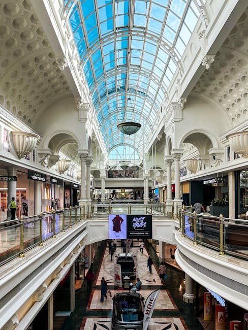 The image depicts the interior of the Bentall Centre shopping mall in Kingston upon Thames, featuring a spacious, multi-level atrium with a large, curved glass roof allowing natural light to illuminate the area. The upper level showcases a row of retail shops with black storefronts and glass display windows, some displaying mannequins and merchandise. The lower level is accessed by a series of escalators, with a few shoppers visible walking through the corridor. The balustrade along the edge of the upper level is made of metal with glass panels, and decorative classical-style wall sconces are mounted on the white, arched walls. The flooring consists of patterned tiles, and digital screens are mounted on the walls, one advertising a clothing sale. This setting provides a neutral backdrop for house removals or furniture transport services, highlighting an environment suitable for moving-related logistics and packing activities during home relocation projects facilitated by removals Kingston upon Thames.