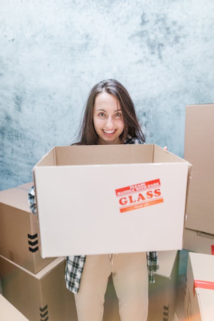 A young woman with long brown hair smiling at the camera while holding an empty cardboard moving box labeled 'Glass' in red, inside a room with a textured blue-gray wall. Around her are several other cardboard boxes, some sealed with tape and others open, with visible packing materials such as crumpled paper and bubble wrap. The scene depicts a home relocation process, with the woman involved in packing or preparing for furniture transport as part of a house removal. The environment suggests an indoor setting, likely a living space or storage area, during the loading or packing stage of a move coordinated by Removals Kingston upon Thames, a professional removals service specializing in house and furniture relocations.