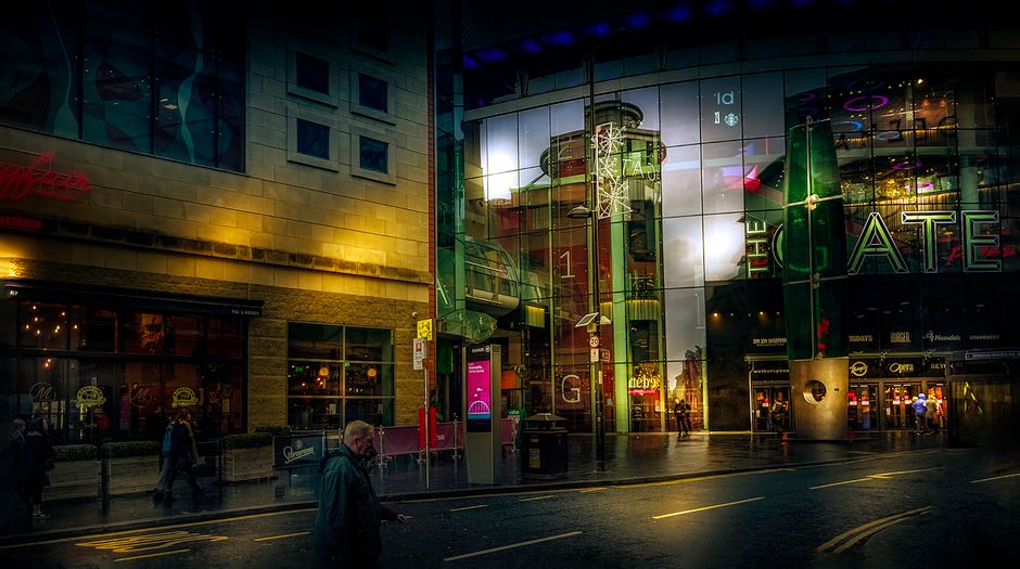 The image depicts the interior of the Bentall Centre shopping mall in Kingston upon Thames, featuring a spacious, multi-level atrium with a large, curved glass roof allowing natural light to illuminate the area. The upper level showcases a row of retail shops with black storefronts and glass display windows, some displaying mannequins and merchandise. The lower level is accessed by a series of escalators, with a few shoppers visible walking through the corridor. The balustrade along the edge of the upper level is made of metal with glass panels, and decorative classical-style wall sconces are mounted on the white, arched walls. The flooring consists of patterned tiles, and digital screens are mounted on the walls, one advertising a clothing sale. This setting provides a neutral backdrop for house removals or furniture transport services, highlighting an environment suitable for moving-related logistics and packing activities during home relocation projects facilitated by removals Kingston upon Thames.