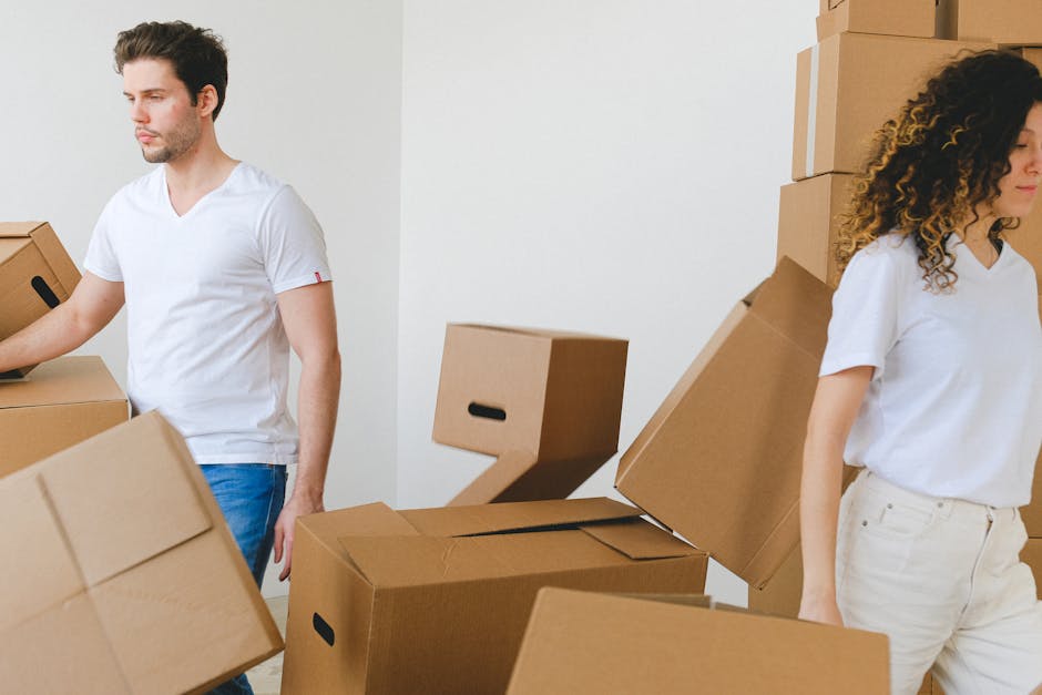 Two individuals, a man and a woman, are engaged in a home relocation process involving packing and furniture transport. The man, wearing a white t-shirt and jeans, is standing on the left side of the image, lifting or adjusting a cardboard moving box. The woman, dressed in a white blouse and light-colored pants, is positioned on the right side, standing beside a stack of cardboard boxes of various sizes, some of which are on the floor and others stacked against a white wall. The boxes are made of brown corrugated cardboard and are sealed with packing tape, indicating they are prepared for moving. The scene appears to be indoors, possibly in a hallway or living room, with natural lighting providing a clear view of the packing and loading process. Visible in the background is a window or a well-lit area, emphasizing an organized effort typical of furniture transport and home relocation. This image exemplifies the typical activities involved when professional removals, such as those offered by Removals Kingston upon Thames, assist with packing, loading, and relocating household items efficiently and carefully.