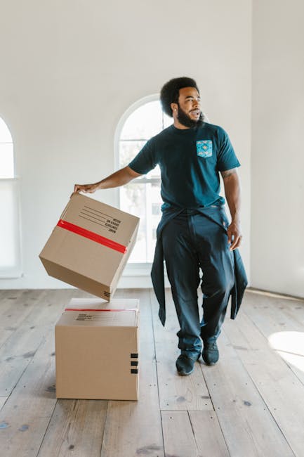 Two individuals, a man and a woman, are engaged in a home relocation process involving packing and furniture transport. The man, wearing a white t-shirt and jeans, is standing on the left side of the image, lifting or adjusting a cardboard moving box. The woman, dressed in a white blouse and light-colored pants, is positioned on the right side, standing beside a stack of cardboard boxes of various sizes, some of which are on the floor and others stacked against a white wall. The boxes are made of brown corrugated cardboard and are sealed with packing tape, indicating they are prepared for moving. The scene appears to be indoors, possibly in a hallway or living room, with natural lighting providing a clear view of the packing and loading process. Visible in the background is a window or a well-lit area, emphasizing an organized effort typical of furniture transport and home relocation. This image exemplifies the typical activities involved when professional removals, such as those offered by Removals Kingston upon Thames, assist with packing, loading, and relocating household items efficiently and carefully.