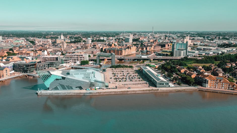 An aerial view of a modern waterfront building with angular architectural design, situated along a river in Kingston upon Thames. The building appears to be part of a busy urban area with a large park