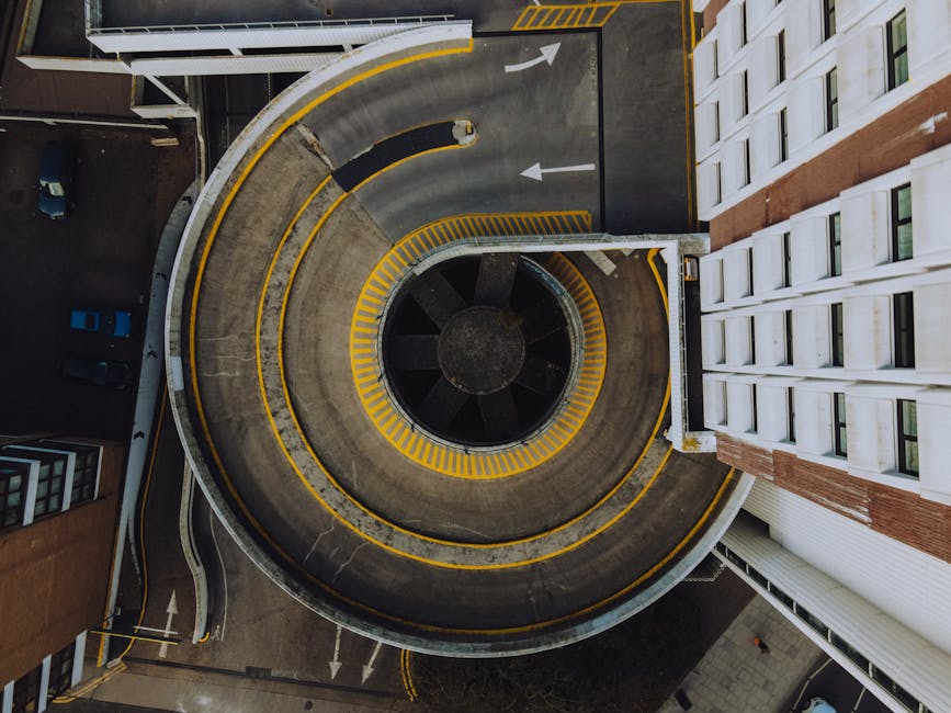 An aerial view of a rooftop parking area on a multi-storey building, with a single silver car parked in one of the spaces near the centre. The parking lot includes designated parking bays marked with