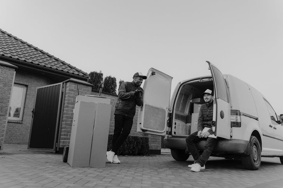Two men are engaged in a home relocation process, loading and managing items outside a residential property. One man is standing on the pavement, wearing a cap, a jacket, and dark trousers, while appearing to be checking a device or planning. The second man is sitting on the open rear of a white moving van, dressed in a hoodie, a hat, and light-colored sneakers, with a smile, indicating a break or communication during the packing and moving process. The van's interior contains cardboard boxes, some wrapped in plastic or fabric materials, indicating packing materials used for furniture transport. Next to the van, there are additional packed boxes and a large black plastic-wrapped item, possibly furniture or appliances, awaiting transport. The setting is outside a house with a tiled roof, brick walls, and a small garden hedge. The environment is illuminated with natural daylight, and the scene captures the loading process as part of a professional furniture transport and moving service provided by Removals Kingston upon Thames.