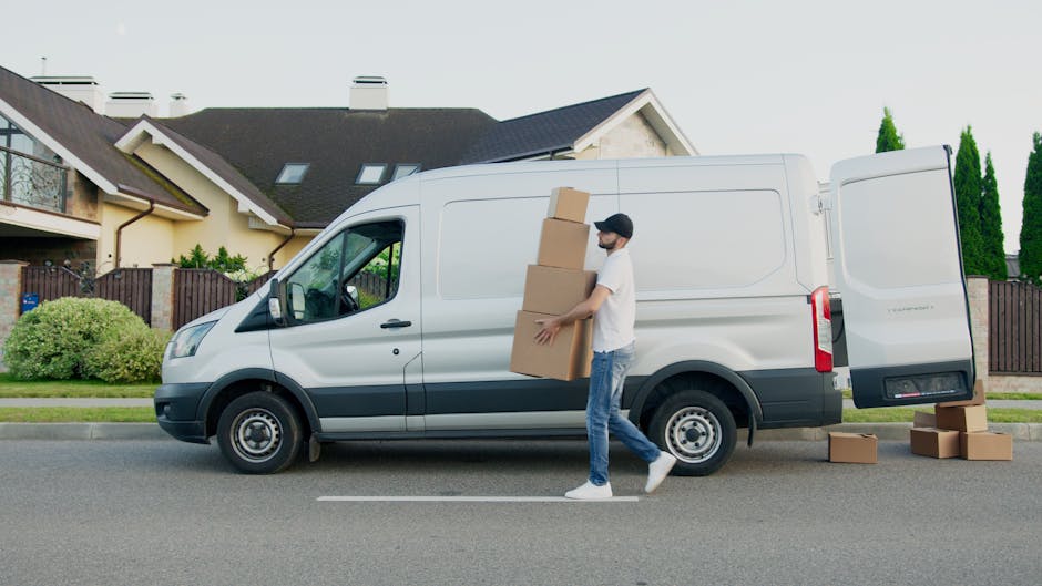 A man wearing casual clothing and a face mask is seen carrying three stacked cardboard boxes towards the open side door of a white delivery van parked on a residential street in Kingston upon Thames. The van, which is part of [COMPANY_NAME]'s home relocation services, is positioned parallel to a driveway leading to a modern, two-storey house with a pitched roof, small windows, and a wooden fence. Several cardboard boxes are placed on the asphalt pavement beside the van, indicating an ongoing packing and moving process. The scene is set during daytime with clear weather, and there are green shrubs and tall trees in the background, contributing to the suburban environment. This image depicts the loading process involved in furniture transport and house removals, emphasizing efficient packing and handling as part of a professional move.