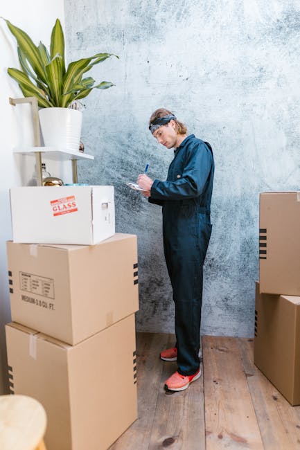 A person wearing protective overalls, a headband, and safety shoes standing inside a room with a textured light blue wall and wooden flooring, surrounded by packed cardboard boxes of various sizes. The individual is holding a clipboard and pen, possibly recording inventory or preparing for a home relocation. Several boxes are stacked on the floor, including one labeled 'GLASS,' indicating fragile items, and others with printed barcodes and shipping labels. To the left, a large potted plant with broad green leaves is positioned on a white shelf, adding a touch of greenery to the space. The scene captures the interior of a property during packing or moving preparations, highlighting careful organisation and handling typical of professional removals, as provided by Removals Kingston upon Thames, serving customer needs in house removals and furniture transport.