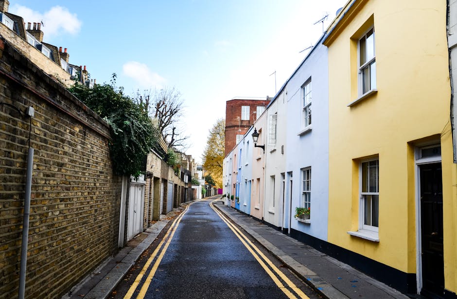 A narrow residential street in Surbiton, Kingston upon Thames, showing a slightly curved tarmac road with double yellow lines along the sides. On the left, there is a tall brick wall with some greenery and small garages or storage units beneath it. On the right, a row of colourful terraced houses painted in pastel shades of yellow, blue, and white, with white-framed sash windows and small flower boxes beneath some windows. The street is wet, suggesting recent rain, and the sky is partly cloudy with patches of blue. In the distance, a red brick building rises above the rooftops, and a few leafless trees are visible, indicating an autumn or winter season. The scene captures an urban environment suitable for home relocation or furniture transport, with houses ready for the loading process, which [COMPANY_NAME] might assist with for house removals on this type of narrow road.