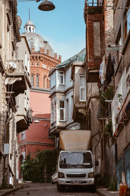 A narrow residential street in Surbiton, Kingston upon Thames, showing a slightly curved tarmac road with double yellow lines along the sides. On the left, there is a tall brick wall with some greenery and small garages or storage units beneath it. On the right, a row of colourful terraced houses painted in pastel shades of yellow, blue, and white, with white-framed sash windows and small flower boxes beneath some windows. The street is wet, suggesting recent rain, and the sky is partly cloudy with patches of blue. In the distance, a red brick building rises above the rooftops, and a few leafless trees are visible, indicating an autumn or winter season. The scene captures an urban environment suitable for home relocation or furniture transport, with houses ready for the loading process, which [COMPANY_NAME] might assist with for house removals on this type of narrow road.